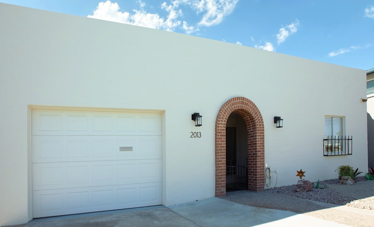 The exterior of the home features a smooth white facade with a welcoming arched entrance. A single-car garage is visible, along with decorative elements such as plants and stones in the surrounding landscape. Clear blue skies and white clouds provide a pleasant backdrop.