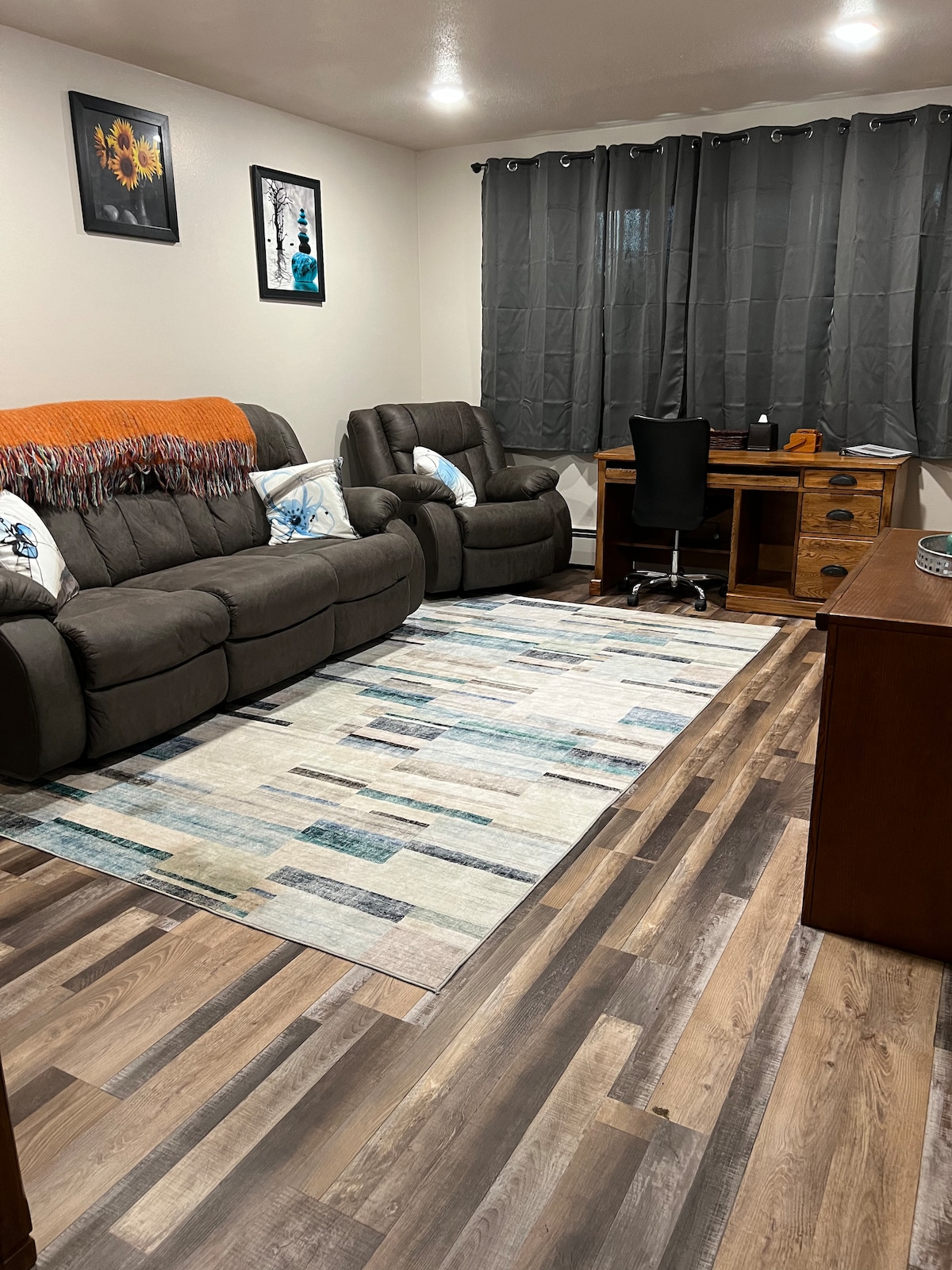 A comfortable living area is presented, featuring a dark gray sofa and armchair, adorned with decorative pillows. A patterned rug lies beneath, complementing the wooden flooring. A desk and chair are positioned against the wall, with curtains providing privacy while allowing light to filter in.