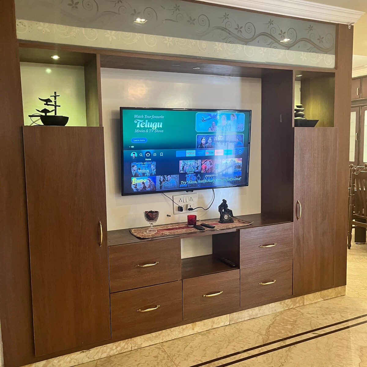 A wooden entertainment unit is showcased, featuring a wall-mounted television. Simple decorative plants are positioned on top. Below the TV, multiple cabinet doors and drawers are arranged for storage, while a small decorative item sits on the surface.