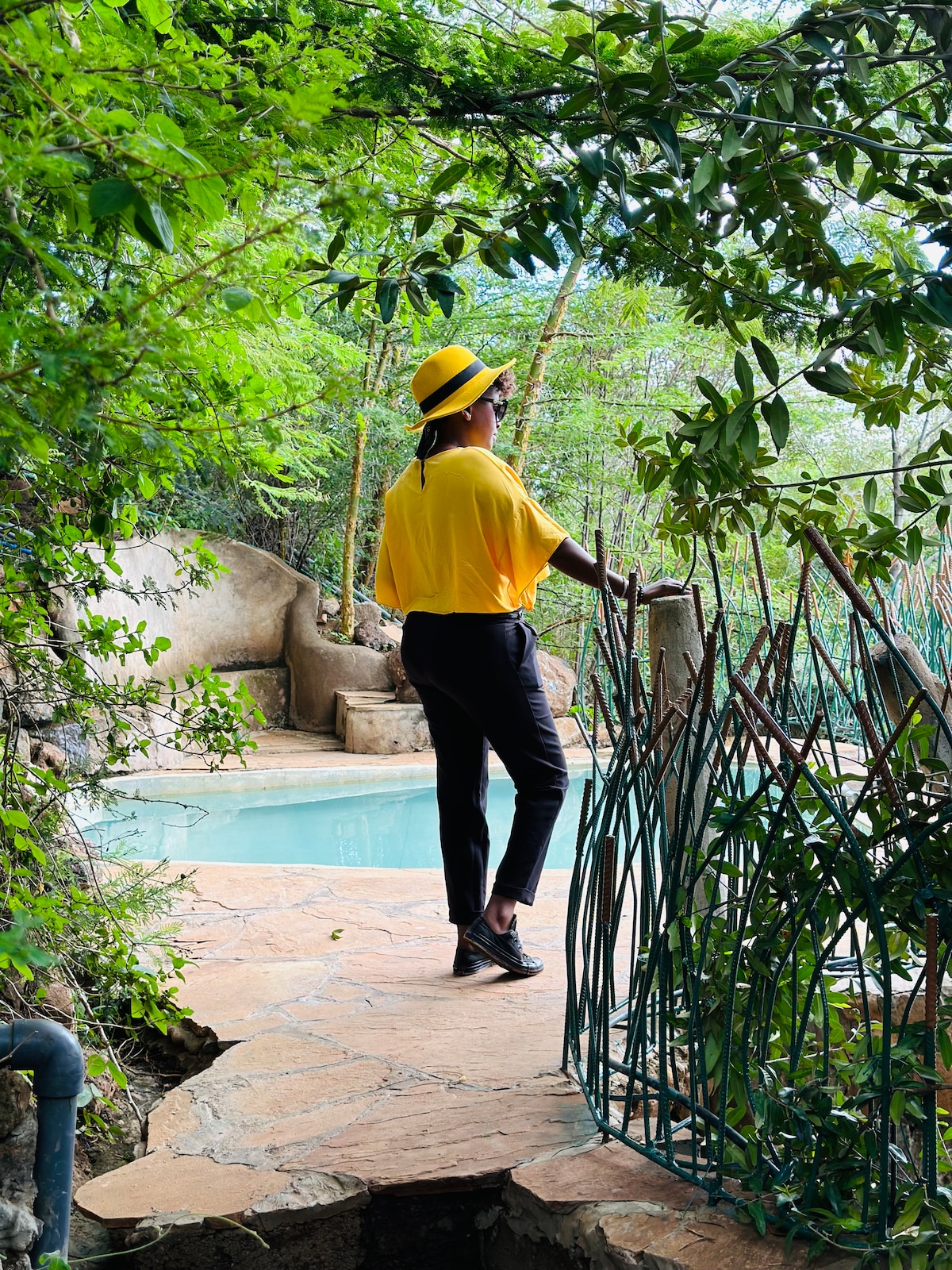 A person wearing a yellow shirt and hat stands on a stone pathway surrounded by lush greenery. To the side, a tranquil swimming pool is partially visible, nestled among natural rock formations. The scene conveys a serene connection with nature.
