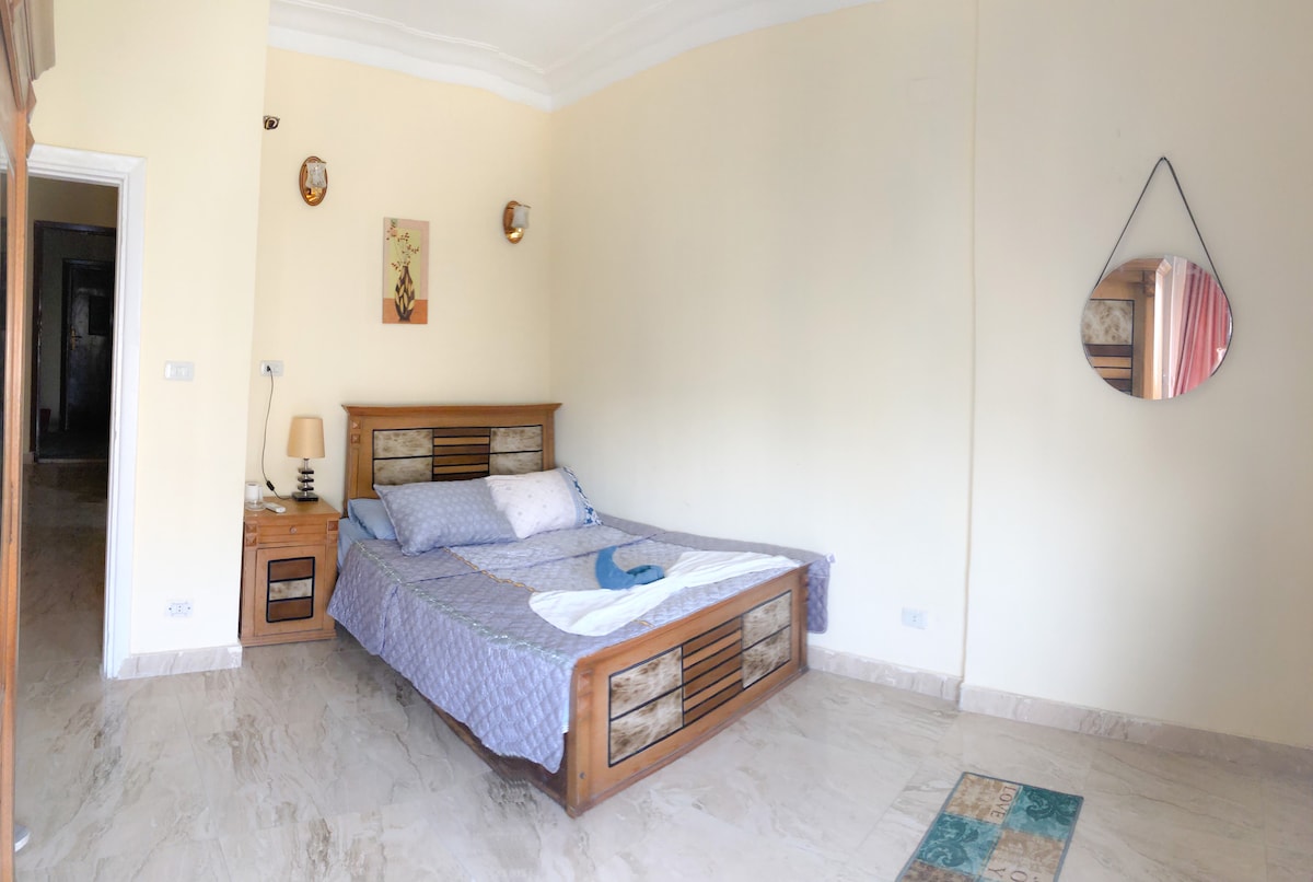 A spacious bedroom is presented, featuring a large bed with a light-colored quilt. A bedside table with a lamp is positioned next to the bed. A mirror is hung on the wall, and natural light filters in through a nearby window.