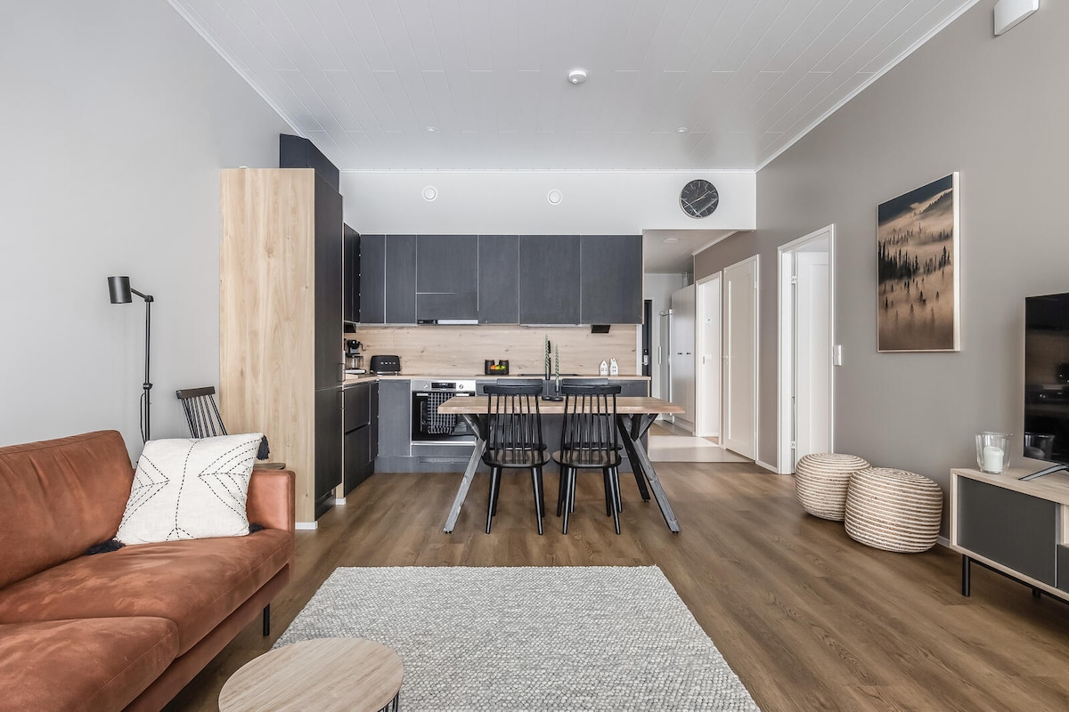 A modern living area is displayed, featuring a brown sofa and a wooden dining table surrounded by black chairs. The kitchen area is equipped with dark cabinetry and contemporary appliances. A light-colored rug anchors the space, while the walls are adorned with soft neutral tones.