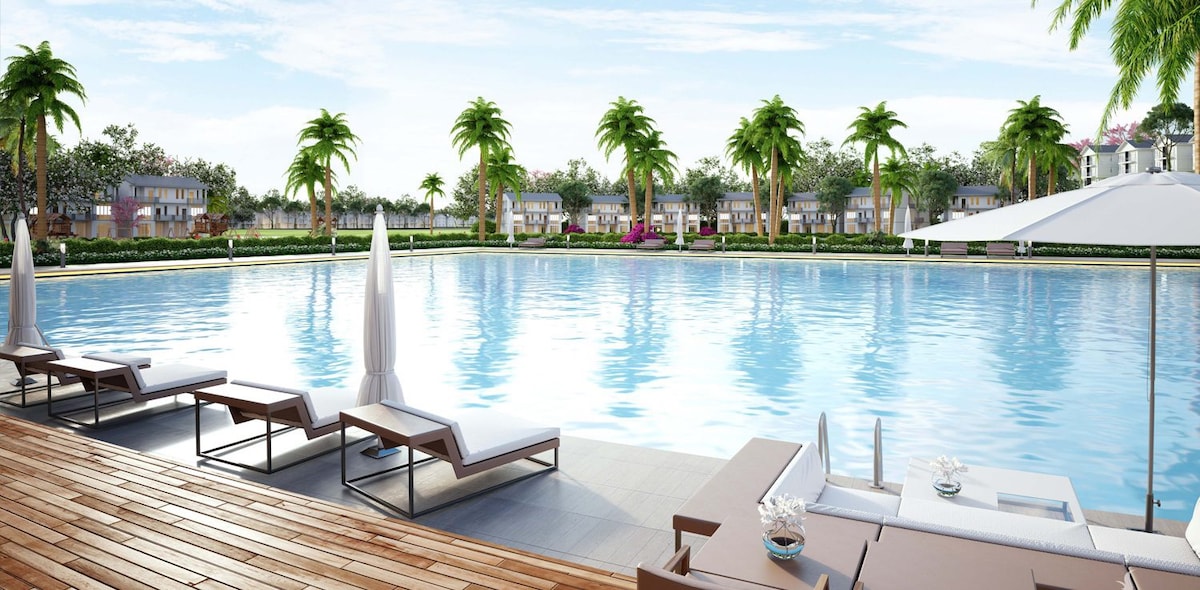 A spacious outdoor pool is surrounded by palm trees, reflecting the clear sky. Comfortable lounge chairs line the pool's edge, offering a relaxed seating area. White umbrellas provide shade, enhancing the tranquil atmosphere of the setting.