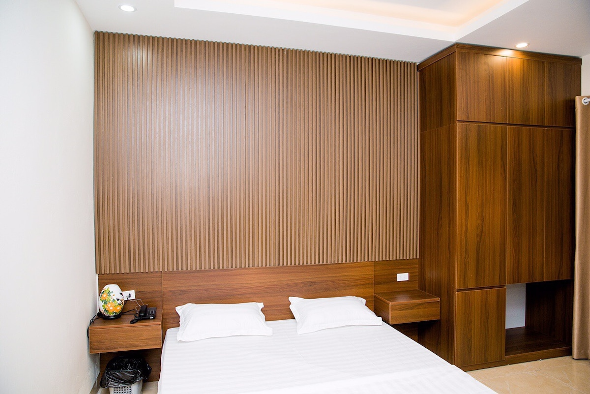 A bedroom featuring a minimalist design with a comfortable bed and a wooden accent wall. A nightstand is positioned beside the bed, and a built-in cabinet is present for storage. Soft lighting illuminates the room, contributing to a calm and restful environment.