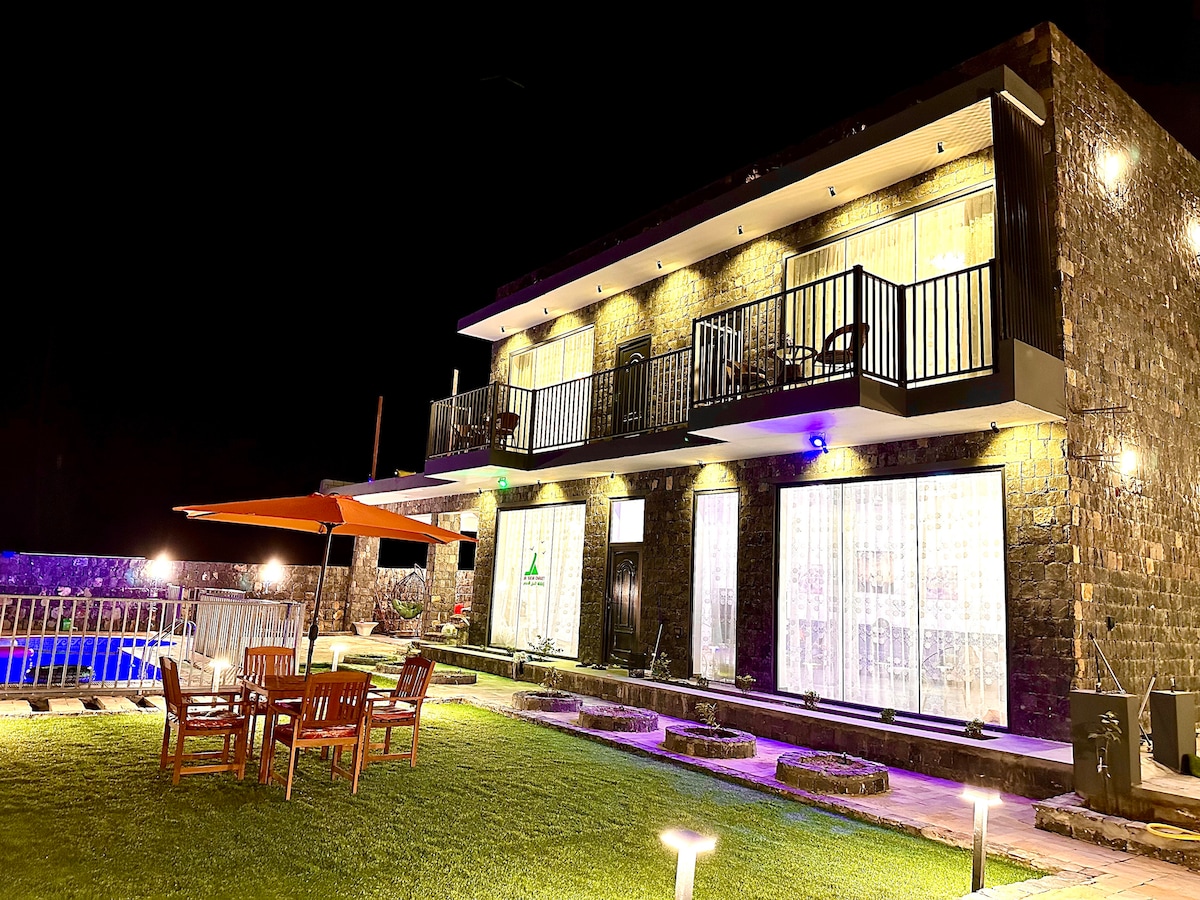 The exterior of the villa showcases a two-story stone façade, illuminated by soft exterior lighting. A patio area features wooden furniture arranged around a table, complemented by a large umbrella. A swimming pool glimmers nearby, adding to the tranquil ambiance.
