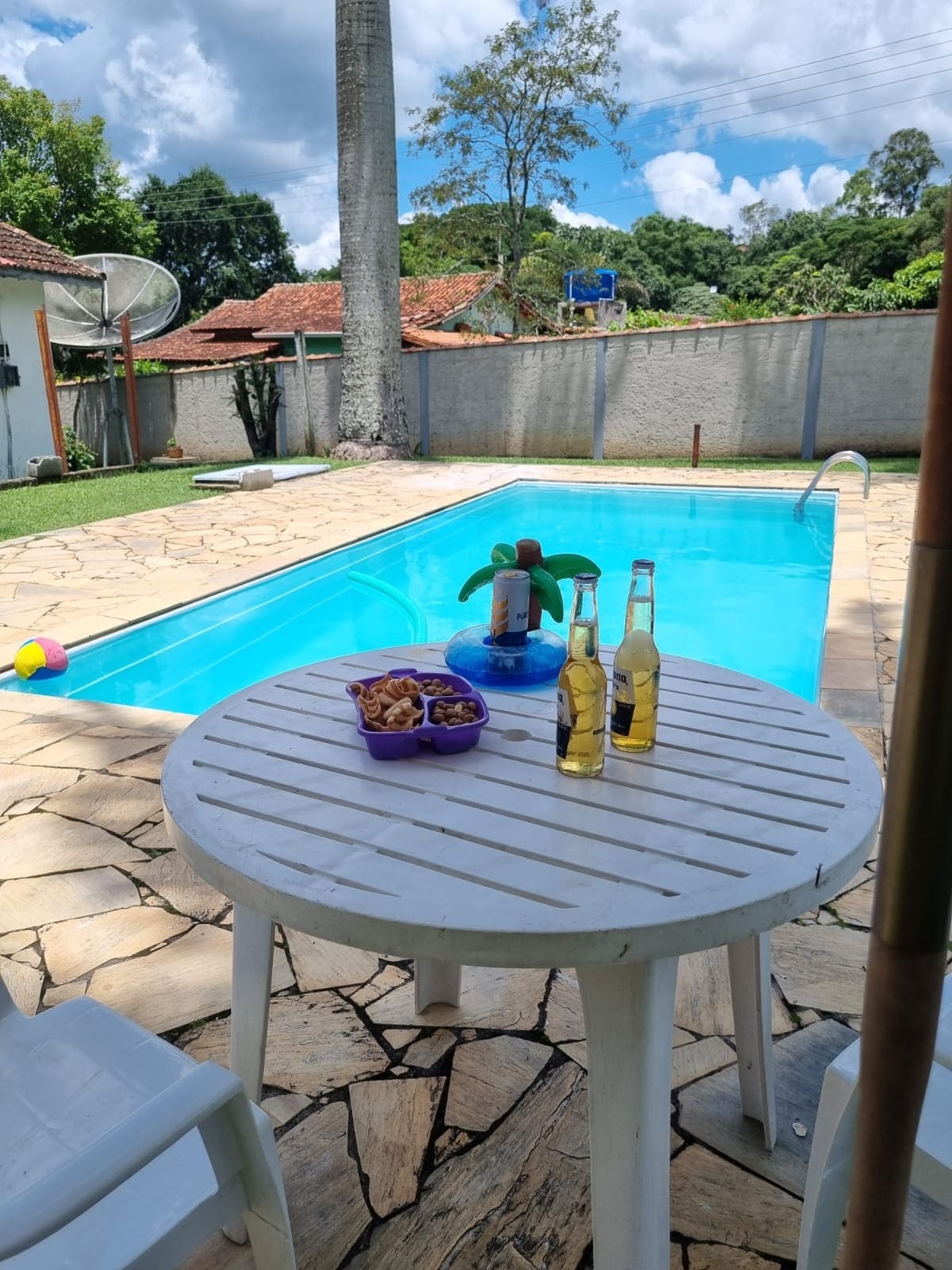 A circular white table is positioned near a clear blue swimming pool, accompanied by two bottles and a container of snacks. The area is surrounded by natural greenery and complemented by a colorful inflatable decoration. A comfortable atmosphere is suggested by the well-maintained outdoor space.