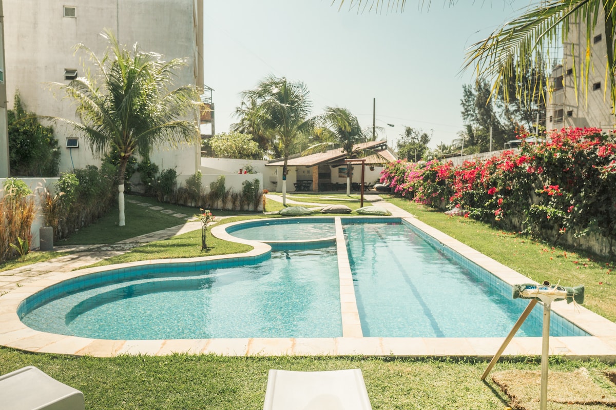 A large swimming pool is set within a landscaped garden, featuring a curved design and surrounded by lush greenery. Tropical palm trees and vibrant flowers are visible along the edges, contributing to a serene outdoor environment ideal for relaxation.