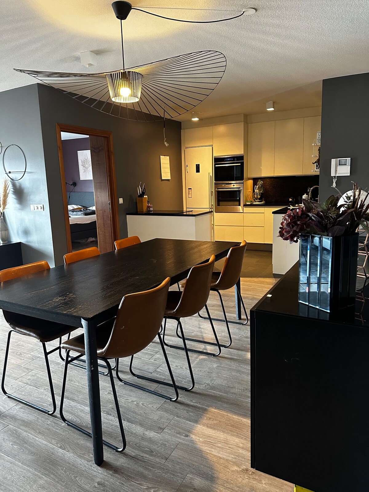 A modern dining area is highlighted with a dark wooden table surrounded by six chairs. The sleek kitchen features light cabinetry and stainless steel appliances in the background. A decorative pendant light hangs above, and a minimalist black cabinet is positioned nearby.