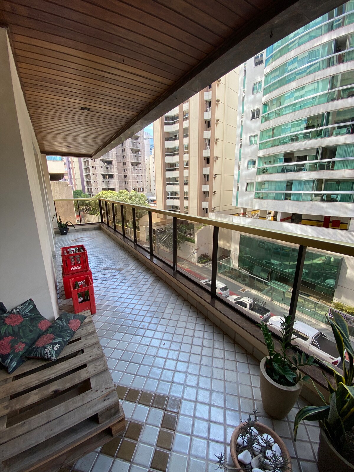 A spacious balcony presents a view of the surrounding buildings and cityscape. The tiled floor features a decorative pattern. A wooden pallet is used as seating alongside vibrant potted plants. Two red chairs are positioned against the railing, offering a spot to relax outdoors.