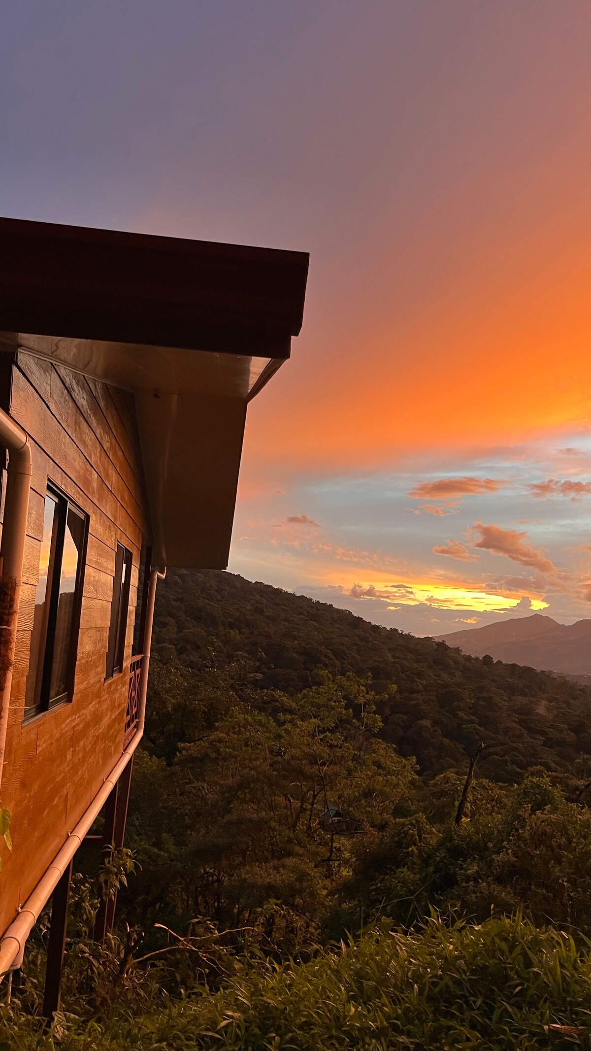 A wooden bungalow is positioned on a hillside, with large windows reflecting the vibrant colors of a sunset. Lush greenery surrounds the property, and distant mountains are visible against a colorful sky.