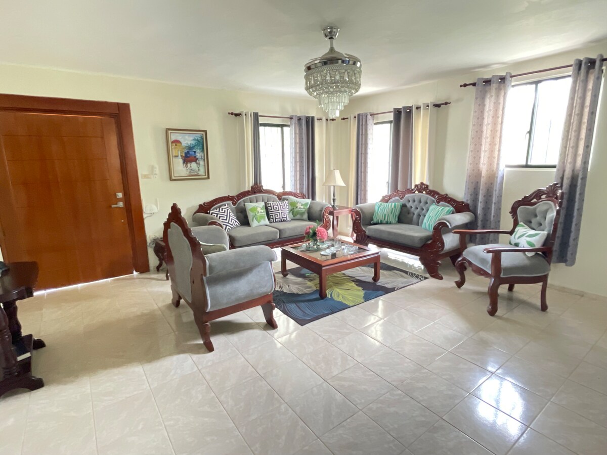 A spacious living room features elegant wooden furniture, including a couch and armchair, arranged around a central coffee table. Large windows allow natural light to fill the space, while patterned curtains add a touch of softness. A decorative rug enhances the inviting atmosphere.