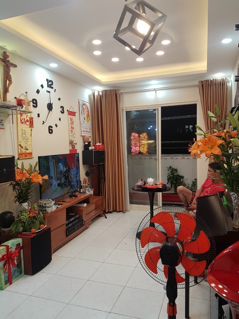 A living area is arranged with a wooden entertainment unit, featuring a television and decorative items. The space is highlighted by a large clock on the wall and lush orange flowers on tables. Soft lighting and curtains enhance the cozy ambiance.