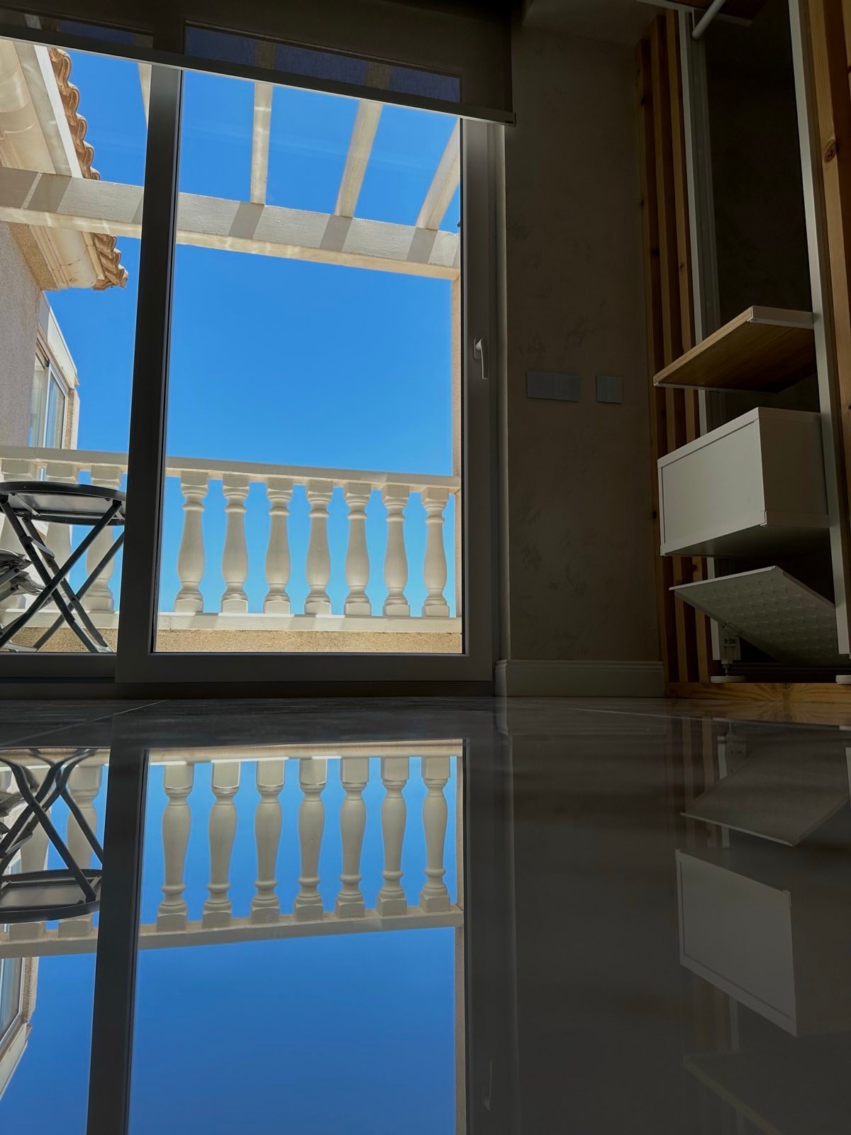 A view is captured through glass doors, revealing a clear blue sky and a balcony with white railings. The reflection of the balcony is visible on the polished floor, enhancing the space's bright and airy feel. A small table and chairs are positioned nearby.