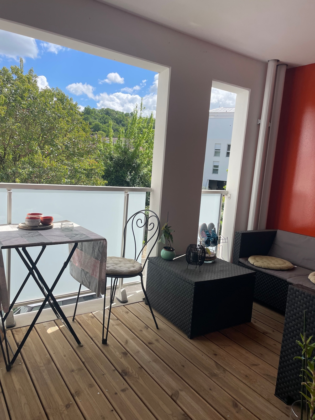 A sunny balcony is presented with a small table and two chairs, offering a relaxing space. A seating area features a dark wicker sofa and a coffee table. Lush greenery is visible through the open windows, enhancing the refreshing outdoor view.