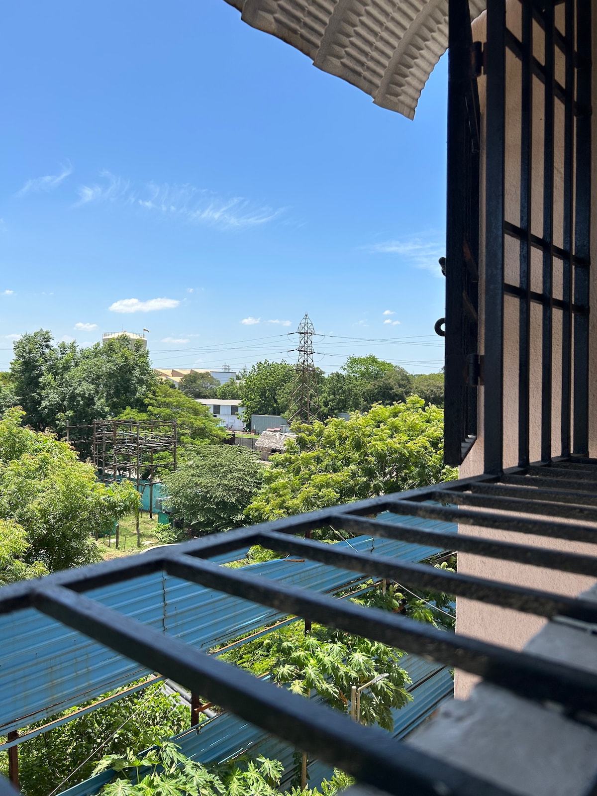 A view from a window frames the greenery outside, featuring trees and buildings that create a lively landscape. Bright blue skies are visible, with occasional clouds passing by. Sunlight casts shadows through the window's bars, enhancing the scene.