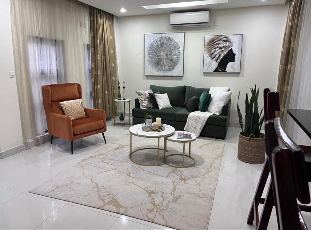 A stylish living area is presented with a modern green sofa and an orange accent chair. Decorative pillows add a touch of comfort, while a marble-patterned rug lies below two round coffee tables. Wall art complements the space, accented by soft, ambient lighting.