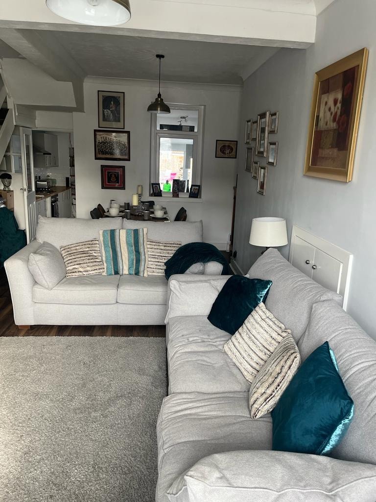 A light-filled living area features two comfortable sofas, one adorned with cushions in shades of teal and muted patterns. A plush area rug lies underfoot, complementing the soft color palette of the walls, while framed artwork adds visual interest to the space.