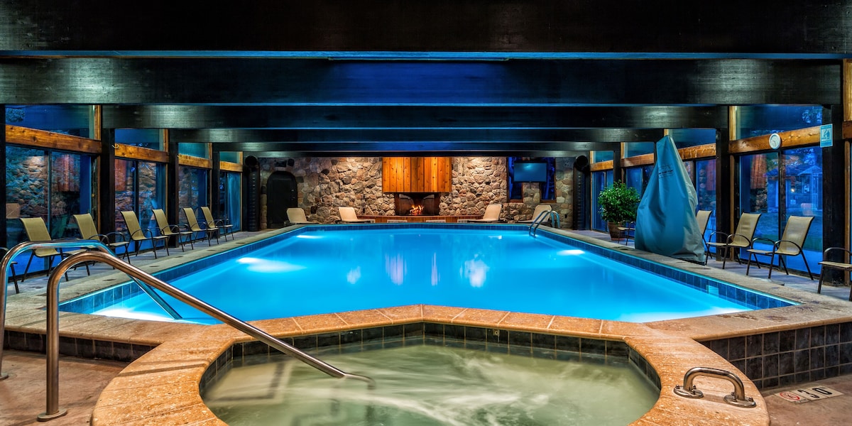 An indoor pool area features a spacious swimming pool surrounded by large windows, providing ample natural light. Lounge chairs line the edges, and a hot tub is positioned to one side. The space is enhanced by warm wood accents and natural stone elements.