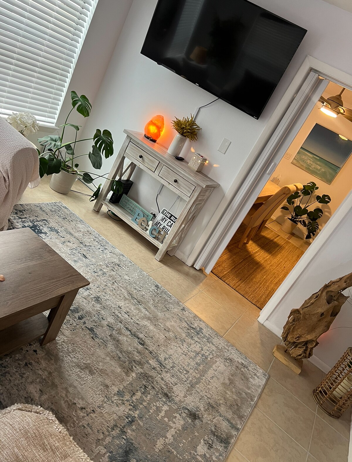 A living area is visible, featuring a light-colored sofa and a wooden coffee table. A large flat-screen TV is mounted on the wall, while a console table holds decorative items and plants. A doorway leads to a well-lit room, enhancing the inviting ambiance.