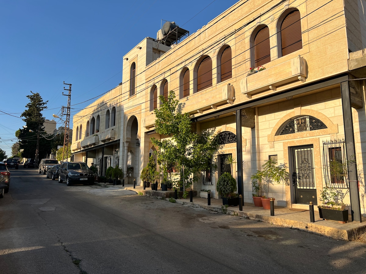 A two-story building is presented, featuring arched windows and a combination of stone and metal accents. Potted plants and shrubs line the pathways. Vehicles are parked along the street, with clear blue skies overhead, reflecting a calm and structured urban environment.