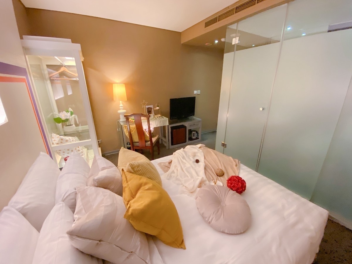A cozy room is presented, featuring a spacious bed adorned with various pillows in neutral and soft colors. A small dining table and two chairs are set near a television. Glass panels provide separation, allowing natural light to fill the space.