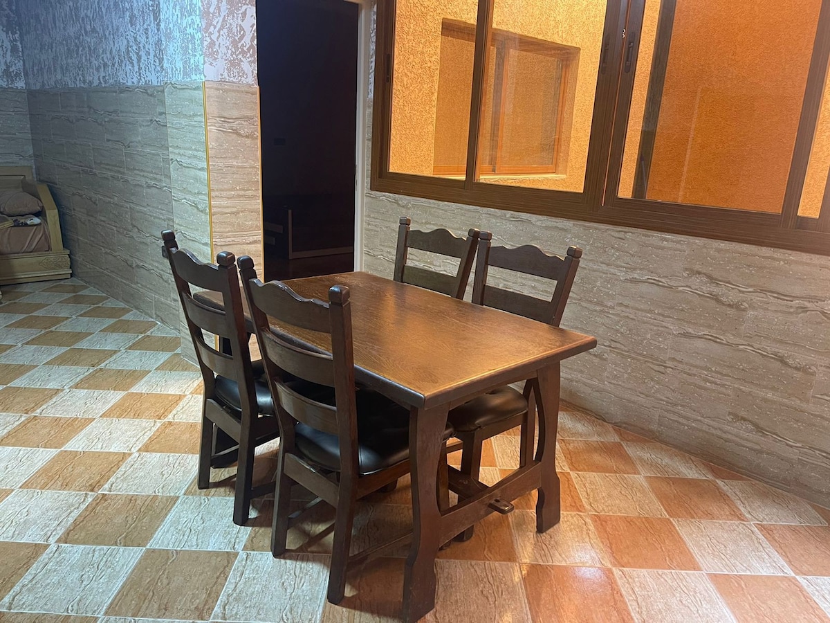 A dining area is showcased with a rectangular wooden table surrounded by four chairs. The floor features a checkered design, and large windows allow natural light to fill the space, highlighting the warm tones of the walls.