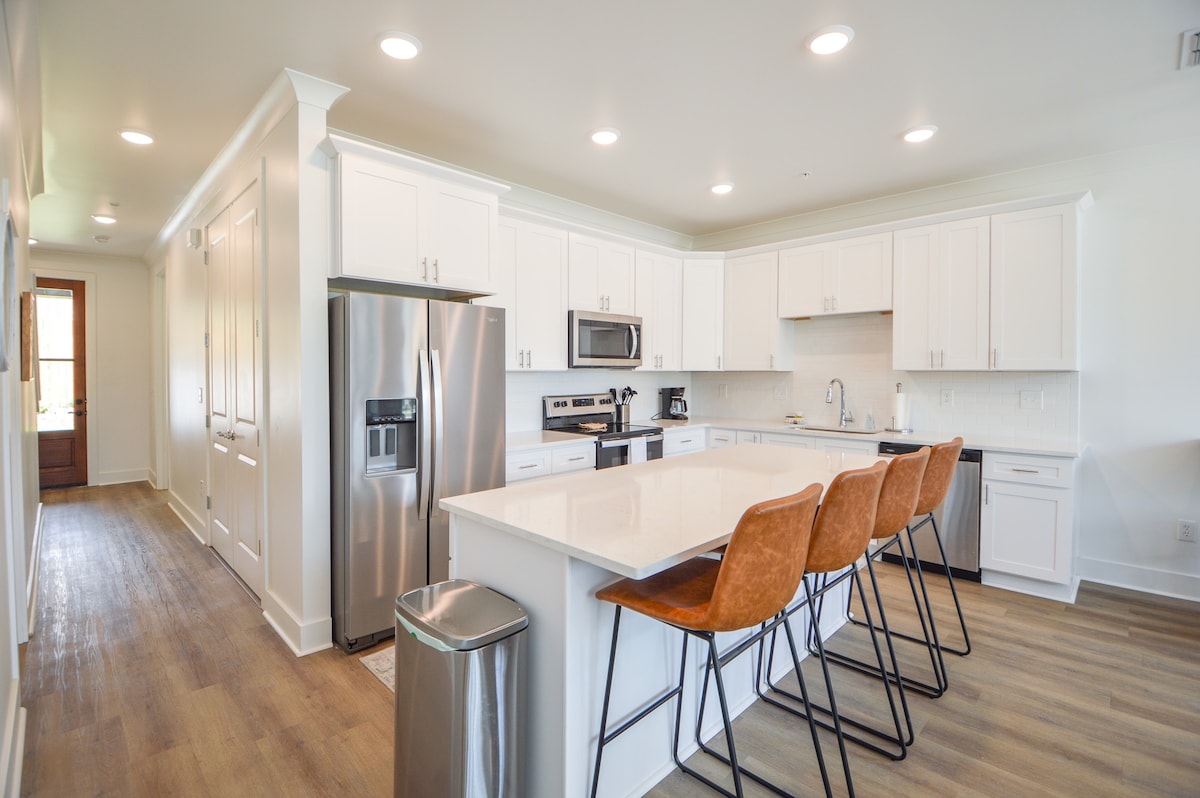 A modern kitchen features white cabinetry and stainless-steel appliances, including a refrigerator and microwave. A central island is equipped with four high-backed bar stools. Natural light fills the space, highlighting the clean lines and open layout.