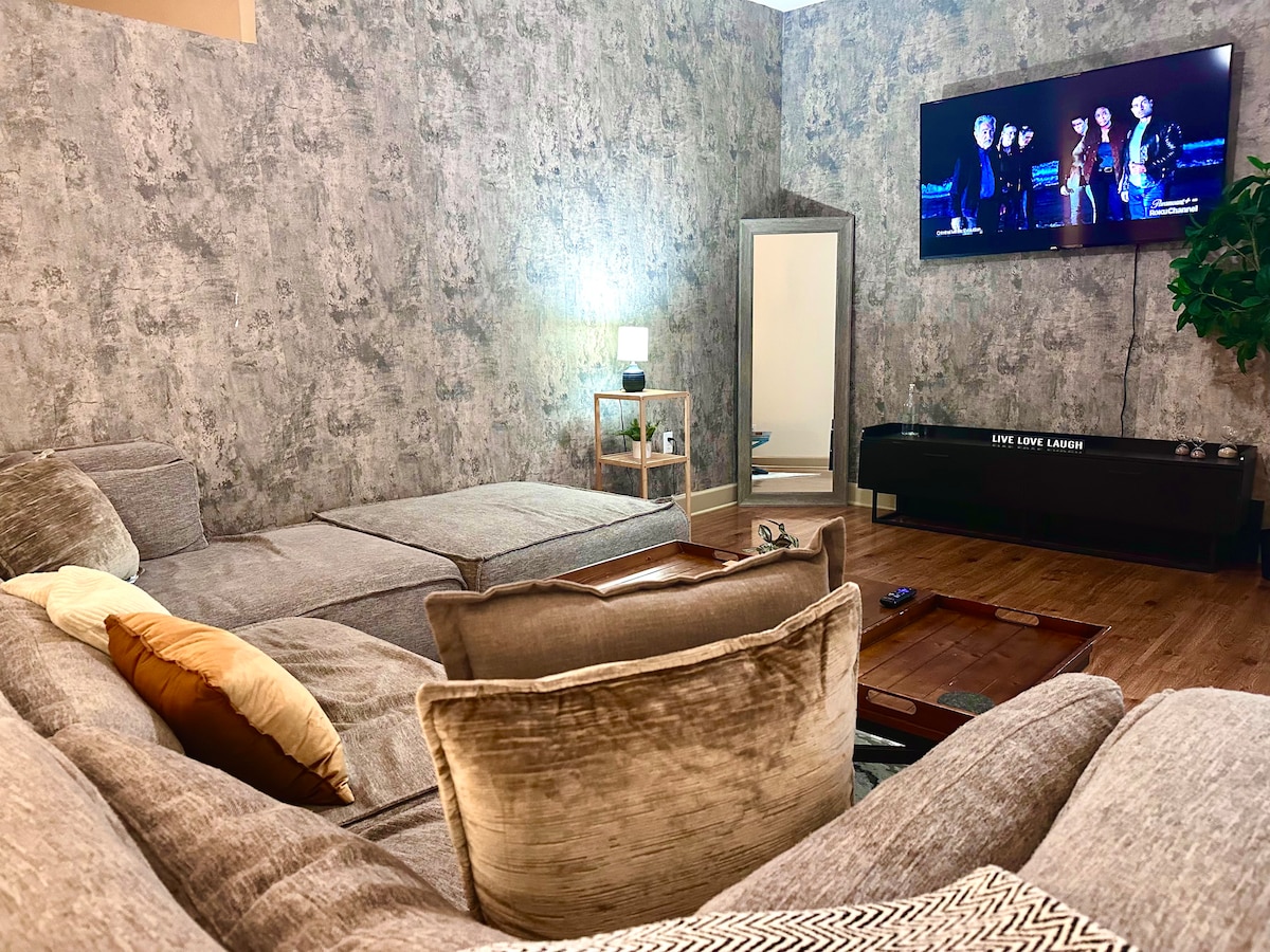 A spacious living area features a large sectional sofa with soft throw pillows, paired with a stylish coffee table. A wall-mounted television displays a group of entertainers. Natural light illuminates the textured wall, while a full-length mirror adds depth to the space.