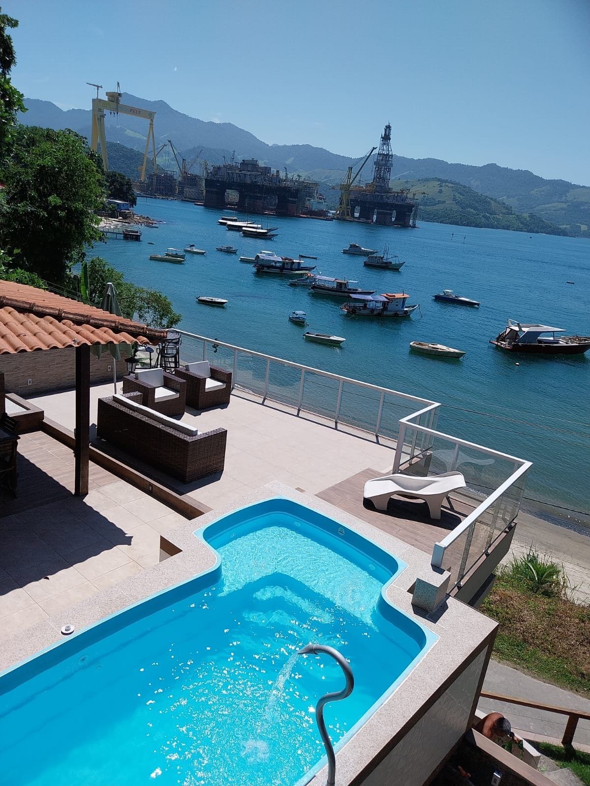 A view of a private swimming pool is presented overlooking a calm waterfront, where various boats are anchored. The outdoor area features a seating arrangement, while mountains and industrial structures create a scenic backdrop against a clear blue sky.