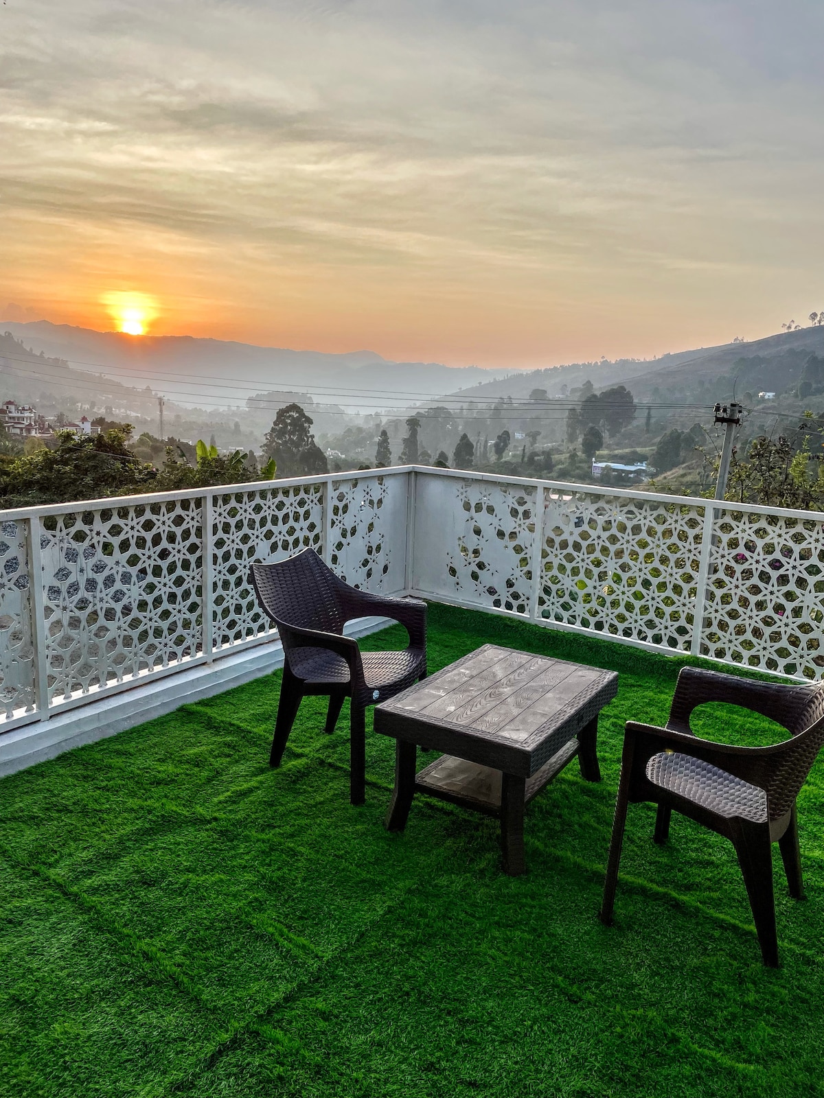 A balcony area is presented with artificial grass flooring, featuring two purple lounge chairs and a low wooden table. The view includes a sunset over rolling hills, with a haze drifting across the landscape, contributing to a serene atmosphere.