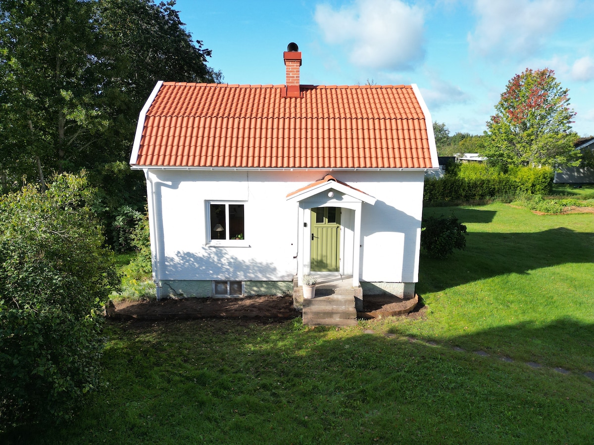 A quaint, white cottage is shown, featuring a distinct red-tiled roof and a green front door. Surrounding greenery and a well-maintained lawn enhance the serene environment. The property is set back slightly from a pathway, providing a private ambiance.