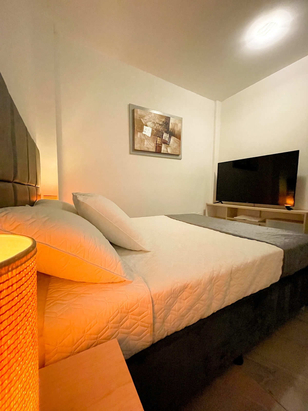 A cozy bedroom features a spacious bed with plush bedding and soft pillows. A framed artwork adorns the wall above, while a flat-screen television is positioned on a wooden shelf. Warm lighting from a nearby lamp creates an inviting atmosphere.
