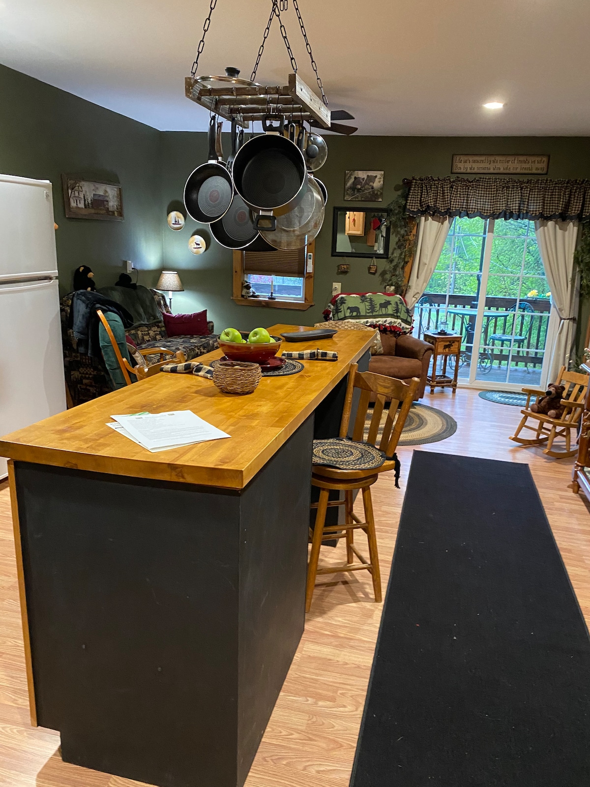 An eat-in kitchen with a wooden breakfast bar is presented, featuring hanging pots above. A white refrigerator and cozy seating area are visible in the background. Natural light enters from sliding glass doors, which lead to a balcony, enhancing the open feel of the space.