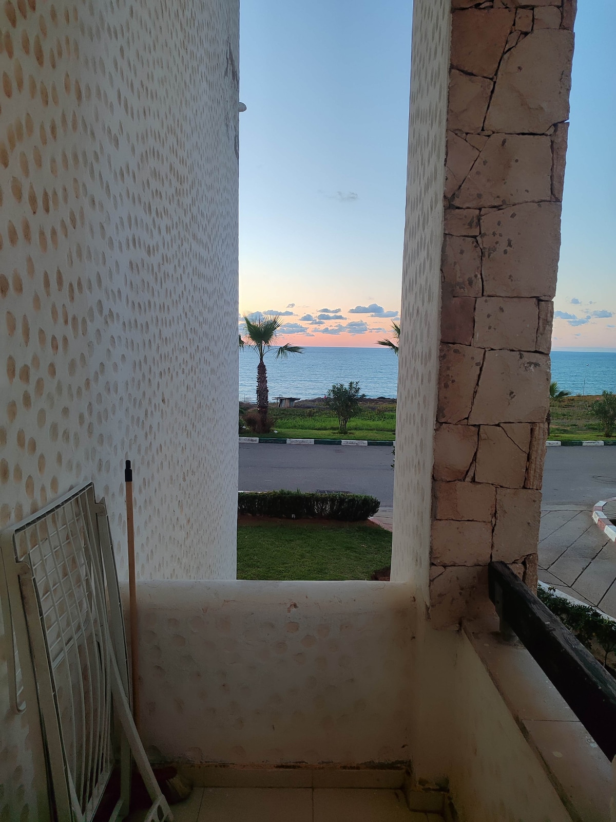 A view from a balcony is portrayed, showcasing the ocean in the distance under a colorful sky at sunset. Palm trees are visible along the shoreline, framed by the textured walls of the building. The floor is tiled and a small railing is present.