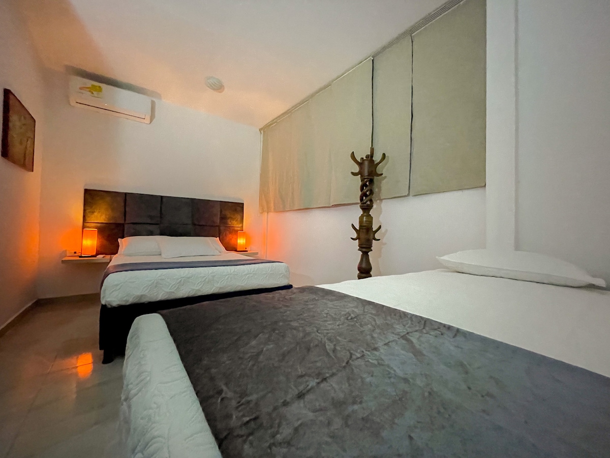 A welcoming bedroom is presented with two spacious beds, each adorned with simple linens. Soft lighting emanates from bedside lamps, creating a calm ambiance. A wooden sculpture stands against the wall, adding a unique decorative touch. Large windows are fitted with neutral blinds.