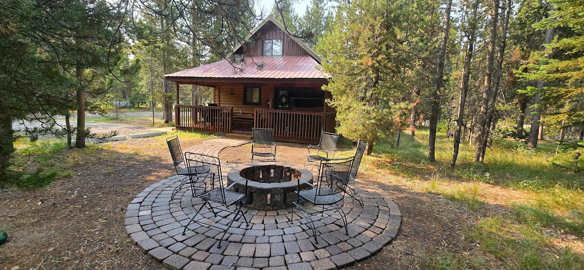 A fire pit surrounded by stone pavers is situated in a natural setting, with several metal chairs arranged around it. The cabin's porch, with a rustic design, is visible in the background, framed by lush trees and gentle landscaping.