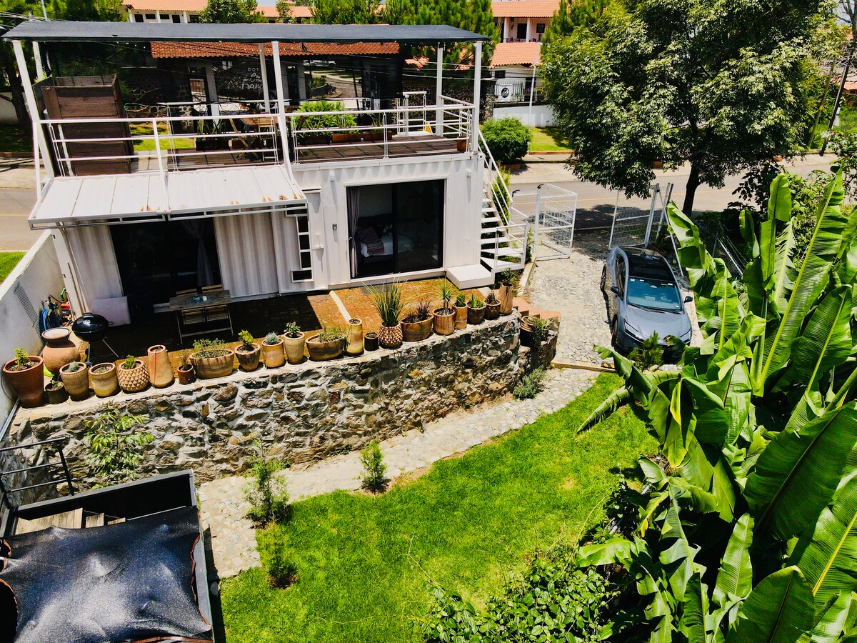 A view of the property reveals a modern structure made from a shipping container, surrounded by greenery. The landscaped yard features a stone pathway and planter pots, while a private car is parked nearby. A spacious terrace overlooks the surroundings, providing an inviting outdoor space.