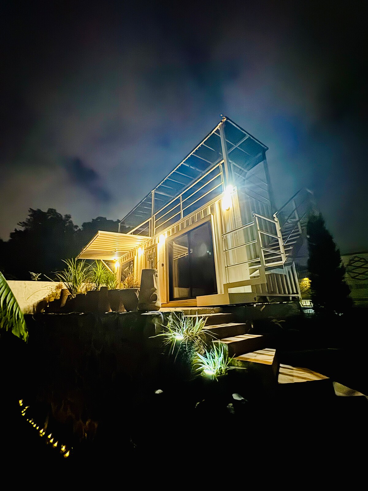 The exterior of the container space is illuminated softly at night, highlighting its modern design. Steps lead up to the entrance, surrounded by low-maintenance greenery and ambient lighting. The upper terrace can be seen, adding to the architectural interest of the structure.
