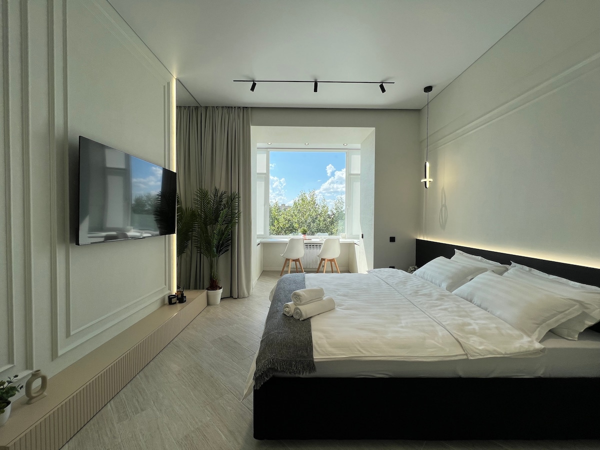 A spacious sleeping area features a large bed dressed in crisp white linens. A television is mounted on the wall opposite, while natural light floods the room through a large window. Two chairs and a small table are positioned by the window, complemented by greenery.
