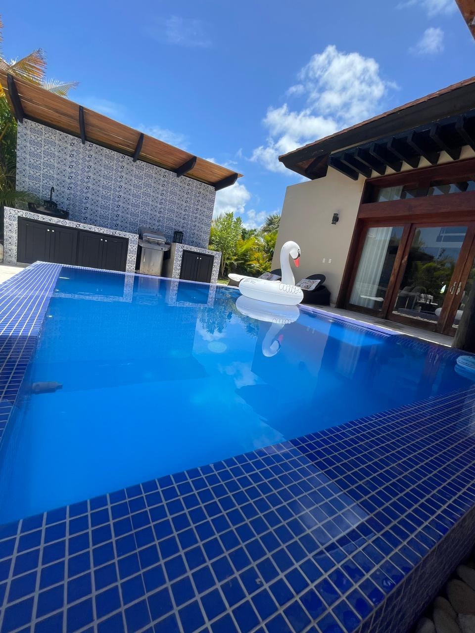 A refreshing pool reflects the vibrant blue sky, with a white swan-shaped float gently resting on the surface. The pool area is bordered by elegant tiles, while a shaded outdoor kitchen can be seen in the background, surrounded by tropical greenery.