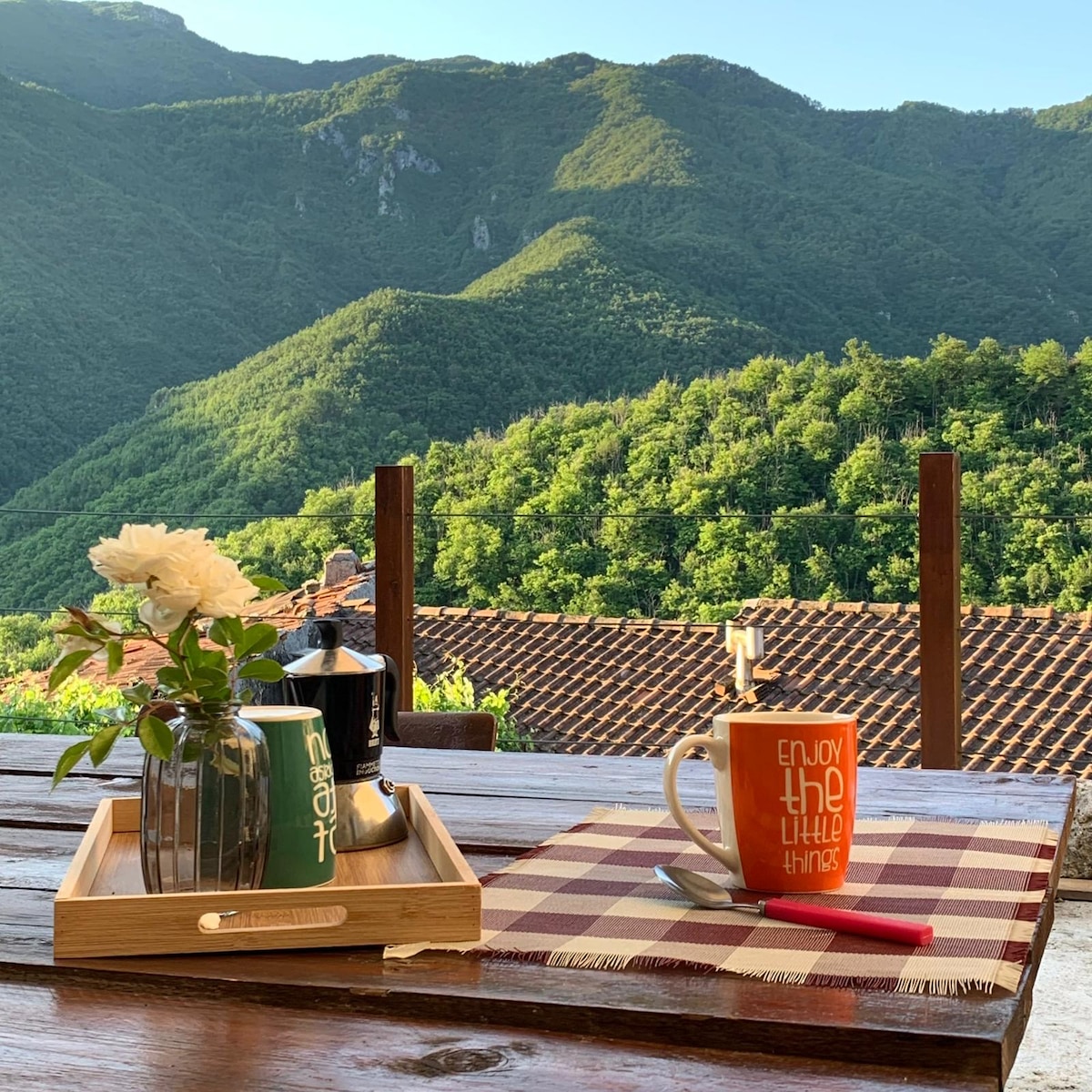 A wooden table is set outdoors with a ceramic coffee mug, a silver coffee maker, and a tray featuring a small bouquet of white flowers. The scene overlooks a lush green hillside dotted with traditional rooftops and framed by the mountains in the background.
