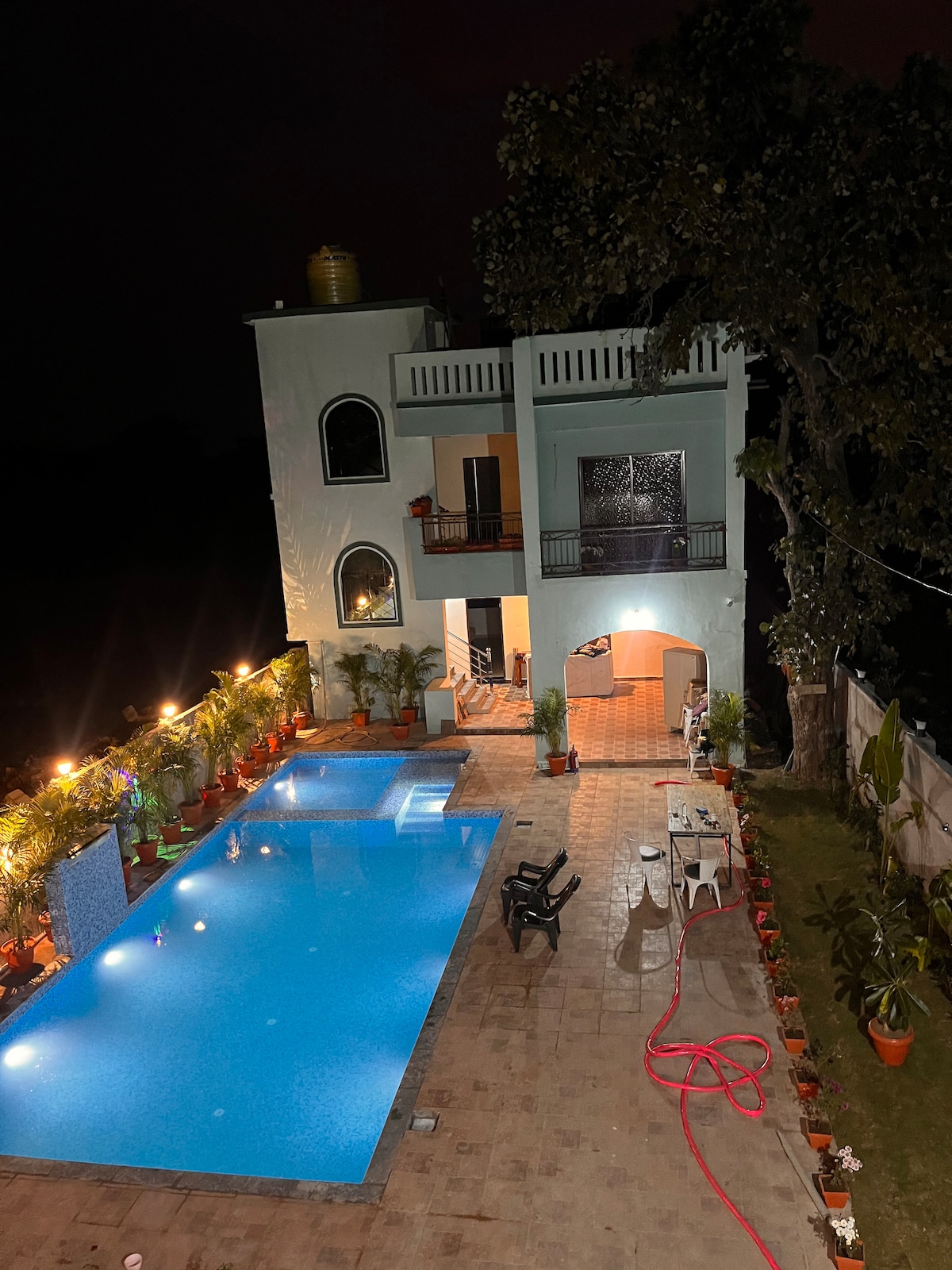 A tranquil pool area is illuminated at night, surrounded by lush greenery and potted plants. The spacious pool is complemented by a nearby building featuring large windows and balconies. A few lounge chairs are positioned beside the pool, enhancing the inviting atmosphere of the space.