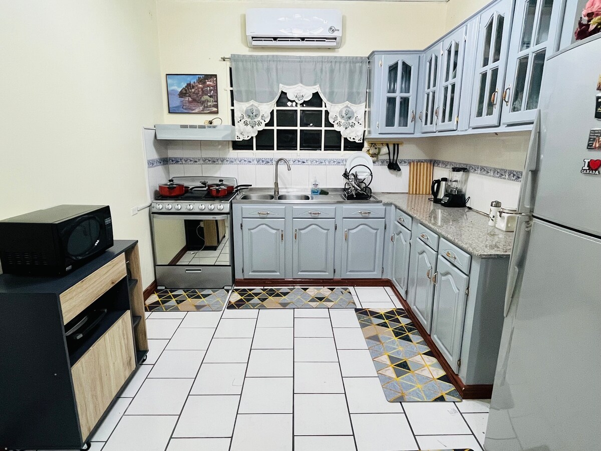 A functional kitchen is highlighted by light blue cabinetry and a granite countertop. A stainless steel stove and refrigerator are present, along with essential appliances and utensils. The tiled floor features a geometric design in yellow and black, enhancing the inviting atmosphere.
