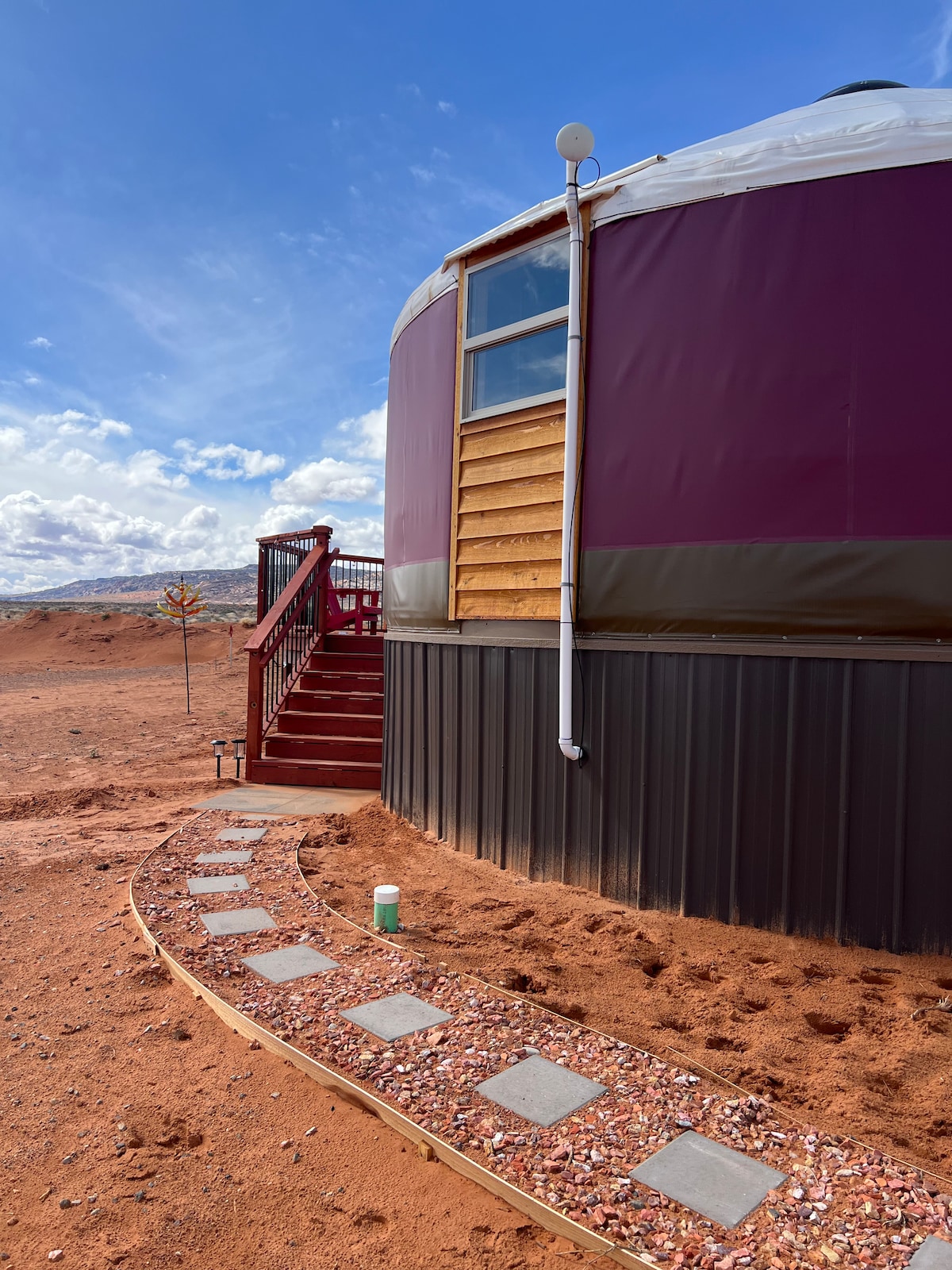 The exterior of a yurt is displayed, featuring a red and brown color scheme. A stone pathway leads to wooden steps, inviting entry. The sky is clear with scattered clouds, highlighting the yurt's structure and surrounding sandy terrain.