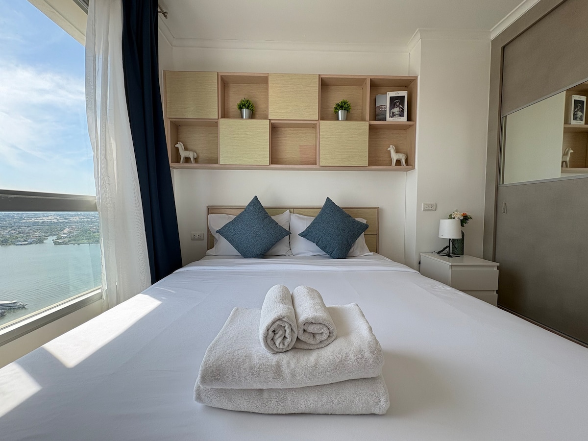 A cozy bedroom features a spacious bed adorned with two blue decorative pillows. Neatly rolled towels are placed on the bed. Natural light enters through large windows, which are framed by light curtains. A set of open shelves above the bed holds various decorative items.
