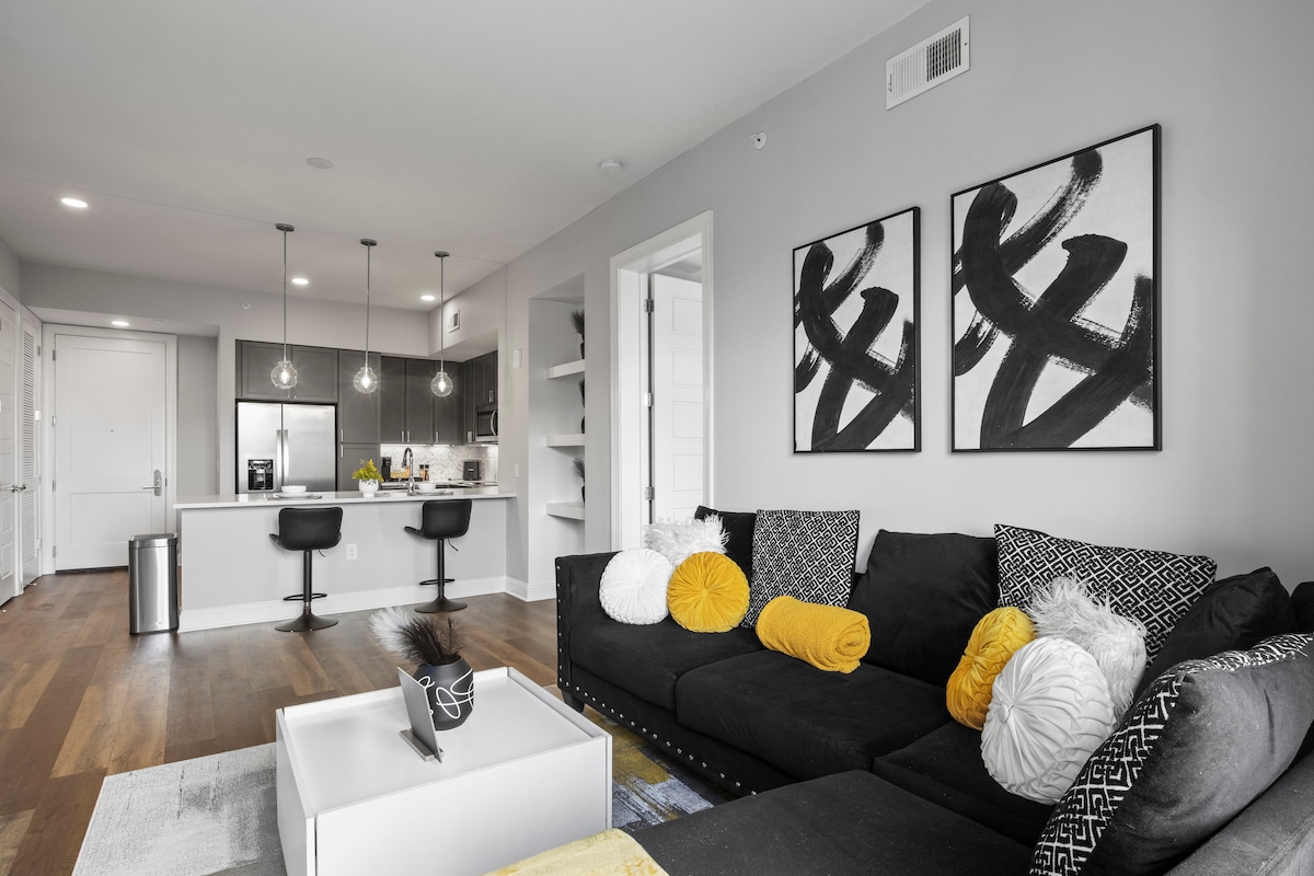 The living area showcases a modern black sectional sofa adorned with decorative pillows in various shades of yellow and white. A minimalist coffee table is positioned in front, while a sleek kitchen with bar seating is visible in the background, enhancing the open floor concept.