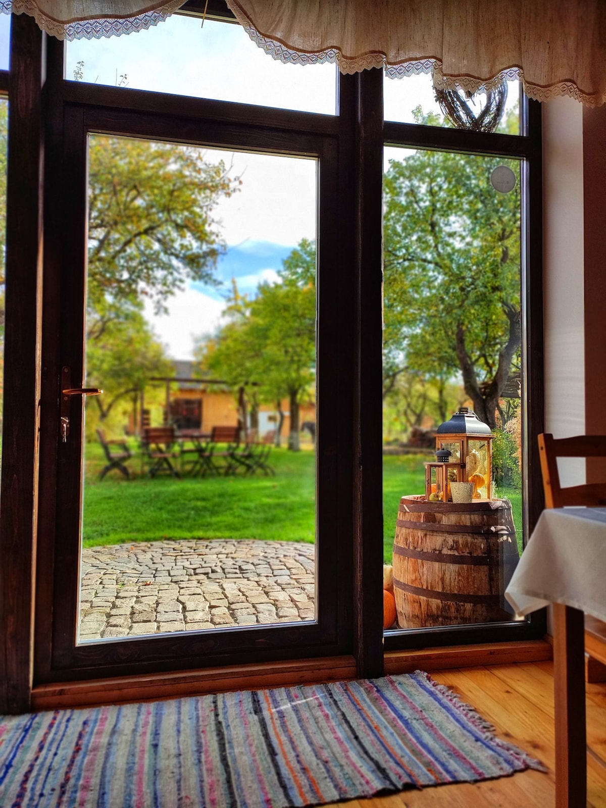 A view through a large glass door reveals a serene outdoor space, featuring a cobblestone path and a grassy area surrounded by trees. A wooden barrel holds a decorative lantern, enhancing the rustic charm of the setting.