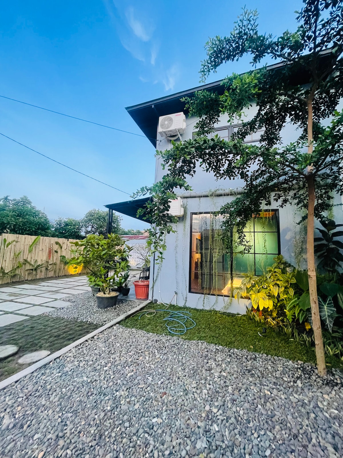 The exterior of the tiny house is shown, featuring a combination of a gravel pathway and a small garden area with greenery. A window with a reflective surface is present, and a climbing plant is visible alongside the building, creating a serene connection with the natural surroundings.