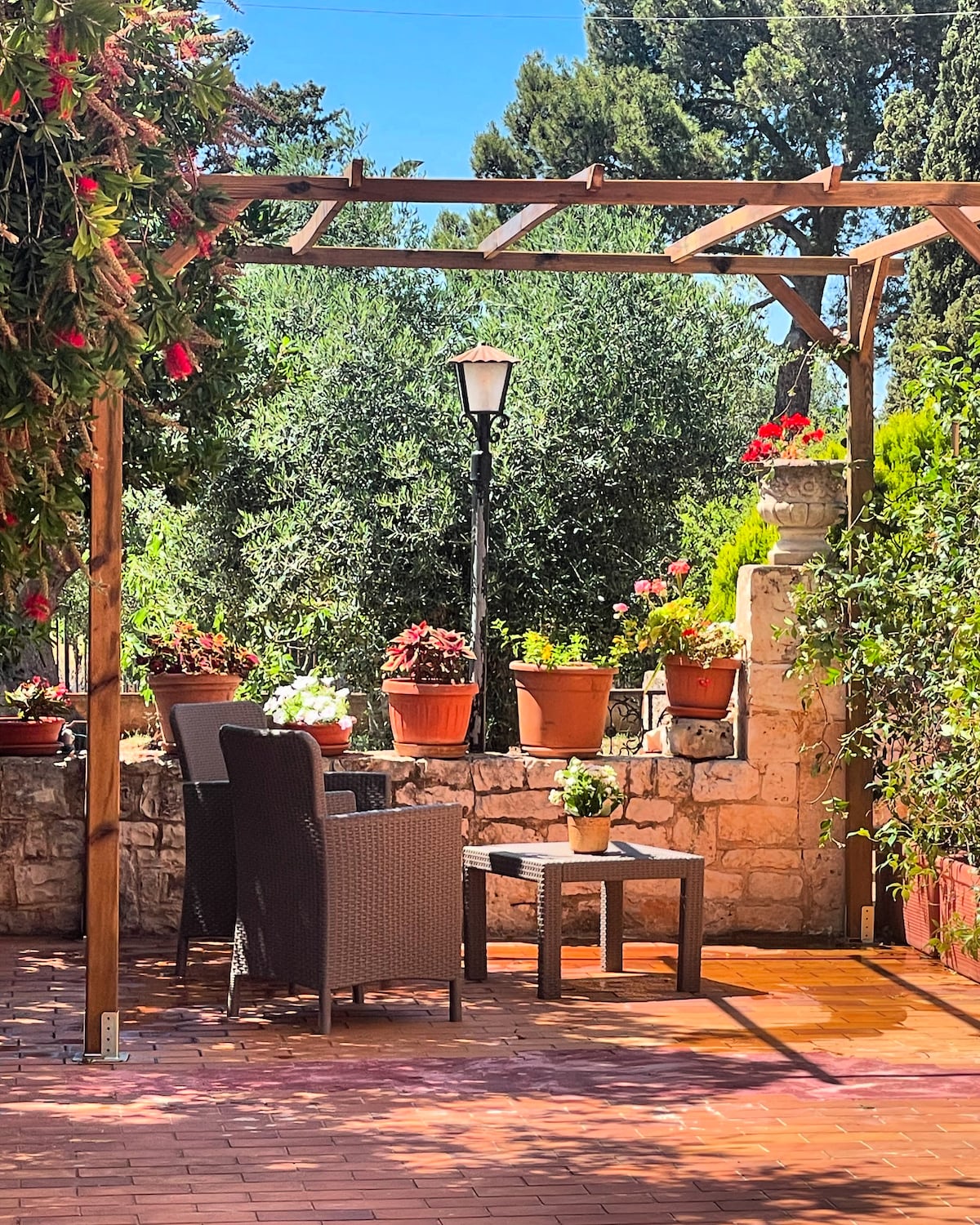 An outdoor seating area is framed by a wooden pergola with climbing plants. Two wicker chairs are positioned beside a small table, surrounded by pots of colorful flowers. A nearby lamppost stands amidst lush greenery, enhancing the serene garden view.