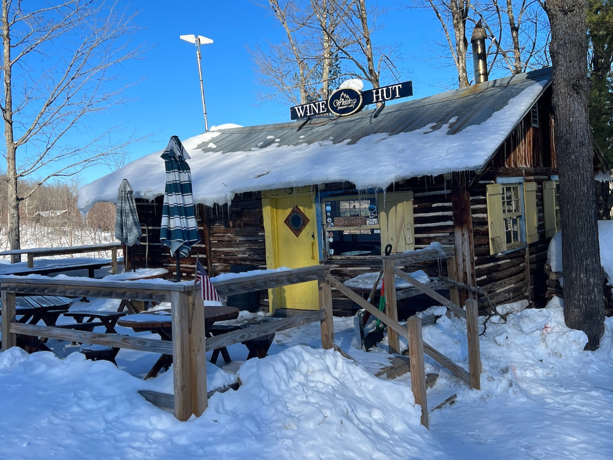 A rustic wooden structure is surrounded by snow, featuring a bright yellow door and a sign reading 'Wine Hut.' Picnic tables are visible on a deck area, partially covered in snow. Icicles hang from the eaves under a clear blue sky.
